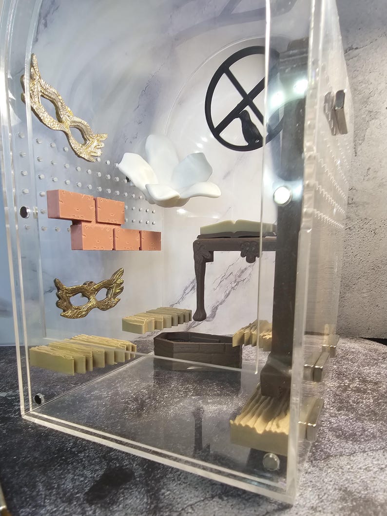 May include: A clear acrylic box with a variety of miniature objects inside, including a white flower, a brick wall, a small table, and two gold masks. The box is sitting on a marble surface.