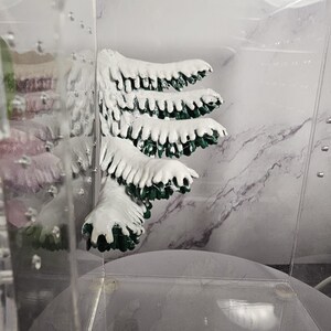 May include: A green and white artificial pine tree branch with a snowy finish. The branch is displayed in a clear glass container.