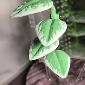 May include: A cluster of three artificial leaves with a light green hue and white edges. The leaves are arranged in a cascading pattern, with a stem connecting them. The leaves are in front of a blurred background.