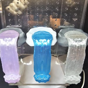 May include: Three clear plastic containers with a white, blue, and purple liquid inside. The containers are on a white surface.