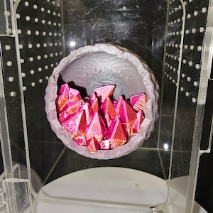 May include: A gray, 3D printed, bowl-shaped object with a textured surface. The bowl is filled with small, pink, iridescent, geometric shapes.
