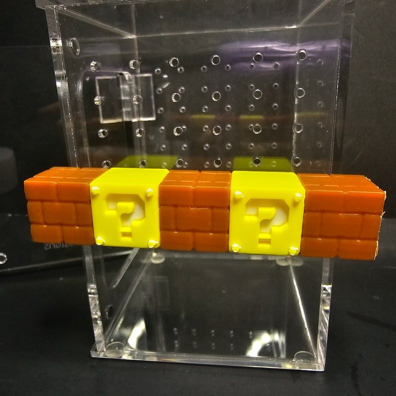 May include: A clear acrylic box with a brown and yellow brick block with a question mark on each block. The brick block is inside the box.