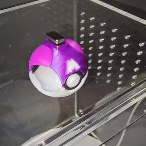 Puede incluir: Una Pok&eacute; Ball morada y blanca con un dise&ntilde;o en blanco y negro. La Pok&eacute; Ball tiene un peque&ntilde;o im&aacute;n plateado en la parte superior.