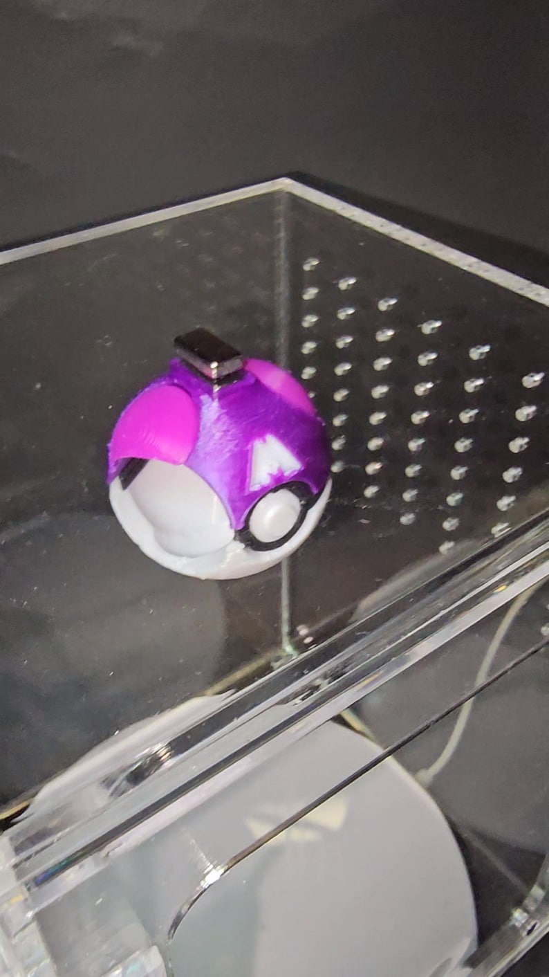 Puede incluir: Una Pok&eacute; Ball morada y blanca con un dise&ntilde;o en blanco y negro. La Pok&eacute; Ball tiene un peque&ntilde;o im&aacute;n plateado en la parte superior.