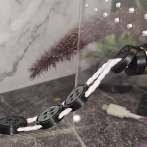 Peut inclure: Une ficelle blanche avec quatre boutons ronds noirs attach&eacute;s. Les boutons sont reli&eacute;s &agrave; la ficelle par une boucle blanche. La ficelle est suspendue &agrave; un objet noir.