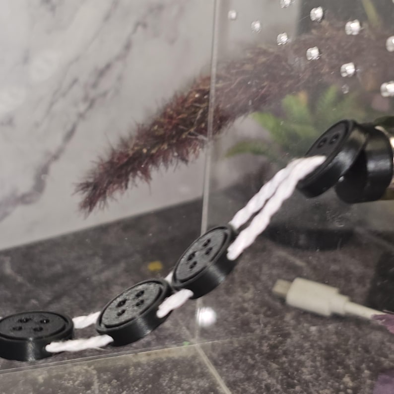 Peut inclure: Une ficelle blanche avec quatre boutons ronds noirs attach&eacute;s. Les boutons sont reli&eacute;s &agrave; la ficelle par une boucle blanche. La ficelle est suspendue &agrave; un objet noir.