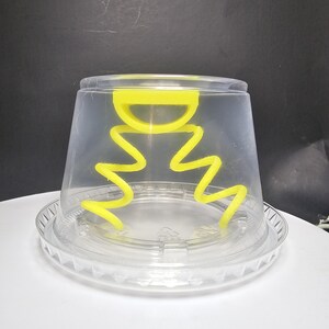 May include: A clear plastic container with a yellow plastic lid. The lid has a handle and a wavy design around the edge.