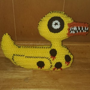 May include: A crocheted yellow duck plush toy with black eyes, a red beak, and black and red accents. The duck has a stitched-on smile and sharp teeth.