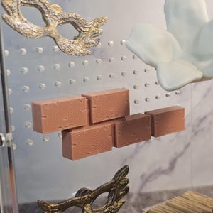 May include: A set of miniature brick pieces in a light brown color, a white flower, and two gold-colored masquerade masks. The items are displayed on a clear acrylic surface with a grid pattern.