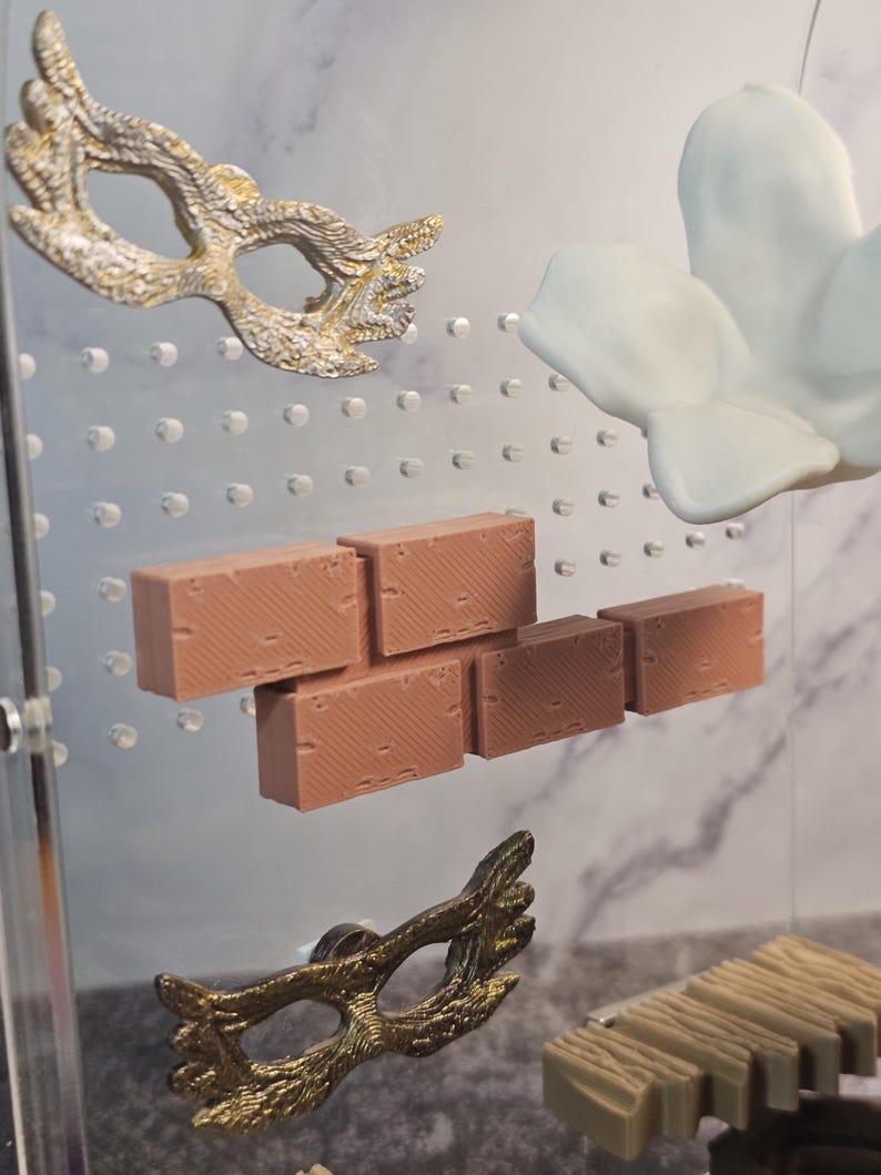 May include: A set of miniature brick pieces in a light brown color, a white flower, and two gold-colored masquerade masks. The items are displayed on a clear acrylic surface with a grid pattern.
