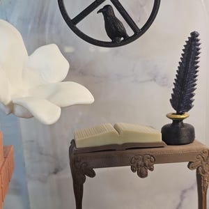 May include: A miniature wooden desk with a closed book on top, a black inkwell with a quill pen, and a white flower in the background.