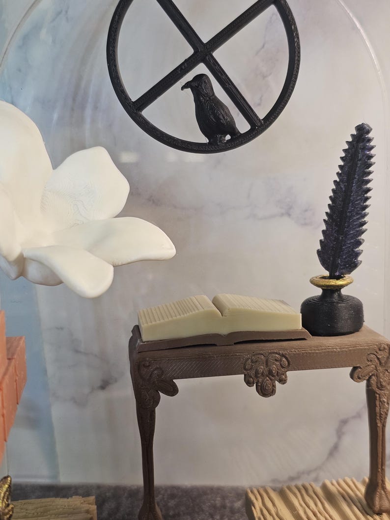 May include: A miniature wooden desk with a closed book on top, a black inkwell with a quill pen, and a white flower in the background.