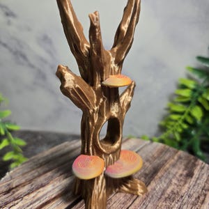 May include: A brown, 3D-printed tree sculpture with three colourful mushrooms. The tree has a textured bark and a hole in the trunk. The mushrooms are orange and pink, with a layered design. The sculpture is on a wooden surface.
