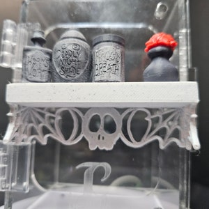 May include: A white shelf with a skull and bat design, holding four small bottles. The bottles are labelled "Worm's Soup", "Deadly Night Shade", "Frog's Breath", and "Poison".