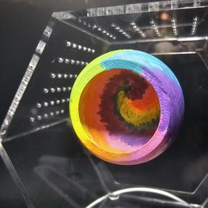 May include: A colorful, rainbow-painted wooden bowl with a spiral design inside. The bowl is displayed in a clear acrylic hexagonal box.
