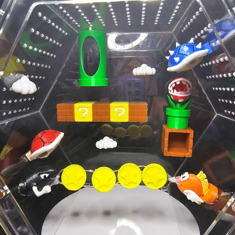 May include: A clear plastic hexagonal box with a variety of colorful 3D printed Super Mario Bros. characters and objects. The characters include a green pipe, a Goomba, a Koopa Troopa, a Piranha Plant, and a Bowser. The objects include a question mark block, a set of coins, and a cloud.