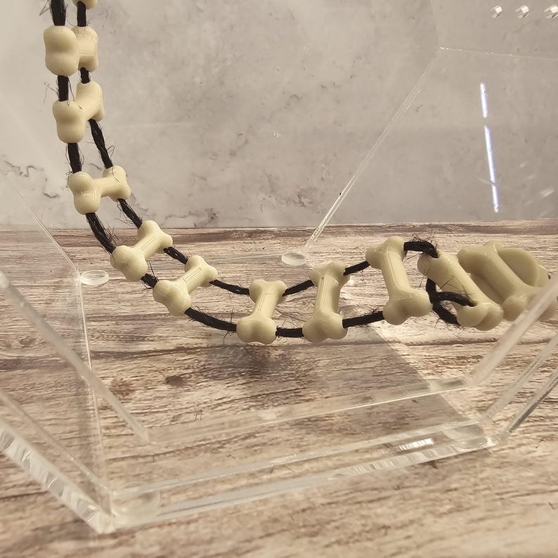 May include: A string of off-white bone-shaped beads strung on a black cord. The beads are arranged in a curved shape and are displayed in a clear acrylic box.