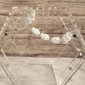 Puede incluir: Una caja hexagonal transparente de acr&iacute;lico con una serie de objetos en forma de nube blanca suspendidos en su interior. La caja est&aacute; abierta por un lado y tiene un fondo de madera.