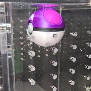 Puede incluir: Un juguete Poke Ball morado y blanco con una banda negra alrededor del medio. El juguete est&aacute; suspendido en un recipiente de acr&iacute;lico transparente con una cuadr&iacute;cula de peque&ntilde;os puntos plateados.