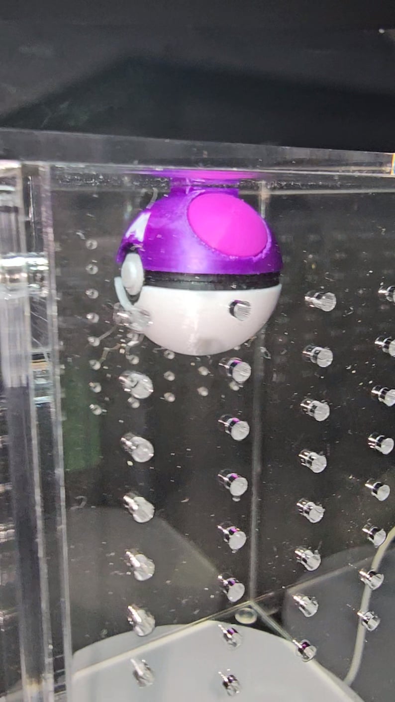 Puede incluir: Un juguete Poke Ball morado y blanco con una banda negra alrededor del medio. El juguete est&aacute; suspendido en un recipiente de acr&iacute;lico transparente con una cuadr&iacute;cula de peque&ntilde;os puntos plateados.