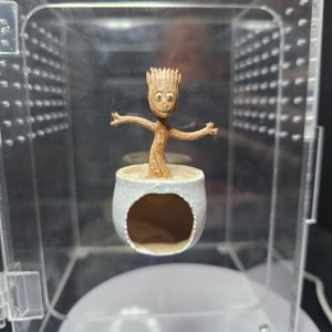 May include: A small, brown, tree-like figure with a smiling face and large eyes stands in a white ceramic pot. The pot has a hole in the side and is suspended in a clear plastic display case.