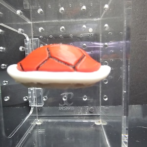 May include: A red and white Super Mario Bros. Koopa shell prop, displayed in a clear acrylic display case with a "JC Designs" logo.
