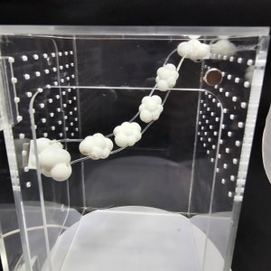 Puede incluir: Una caja de acr&iacute;lico transparente con una cuerda blanca de peque&ntilde;os objetos redondos en forma de nube en su interior. Los objetos est&aacute;n enhebrados y se mueven en un movimiento circular.