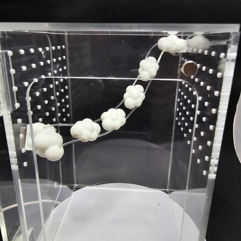 Puede incluir: Una caja de acr&iacute;lico transparente con una cuerda blanca de peque&ntilde;os objetos redondos en forma de nube en su interior. Los objetos est&aacute;n enhebrados y se mueven en un movimiento circular.