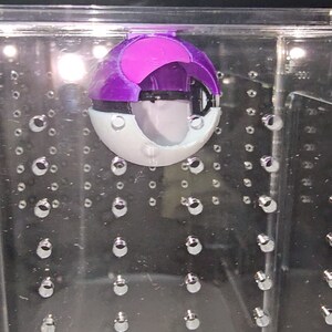Puede incluir: Un juguete Poke Ball morado y blanco dentro de una vitrina de acr&iacute;lico transparente con una cuadr&iacute;cula de peque&ntilde;os puntos plateados.