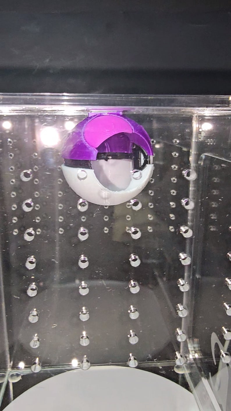 Puede incluir: Un juguete Poke Ball morado y blanco dentro de una vitrina de acr&iacute;lico transparente con una cuadr&iacute;cula de peque&ntilde;os puntos plateados.