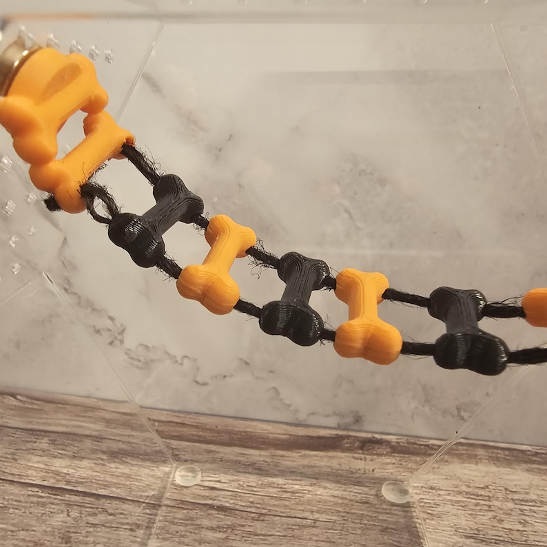 May include: A black and orange dog bone chew toy chain. The chain is made of alternating black and orange bone shapes connected by black string.