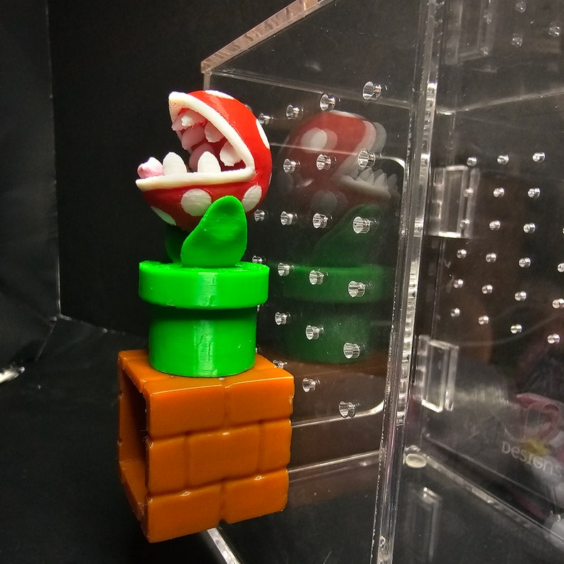 May include: A 3D printed red and white Piranha Plant from the Super Mario Bros. video game series. The plant is in a green pipe and a brown brick base.