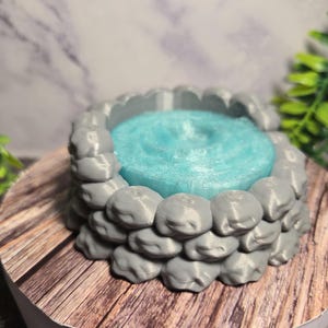 May include: A 3D-printed miniature well with a gray stone exterior and a light blue interior. The well is round and sits on a wooden surface, with a blurred background.