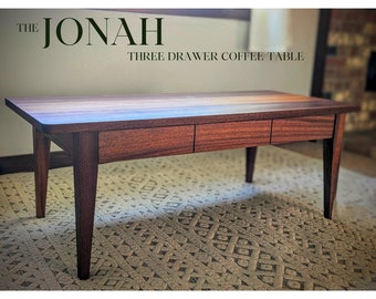 May include: A dark wood coffee table with three drawers. The table has a rectangular top and four legs. The table is called "The Jonah Three Drawer Coffee Table".