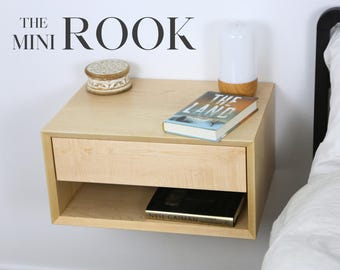 Pode incluir: Uma mesa de cabeceira flutuante de madeira clara com uma gaveta e uma prateleira. A mesa de cabeceira tem um livro intitulado "The Land" e uma pequena caixa decorativa no topo. O texto "THE MINI ROOK" é visível no fundo.