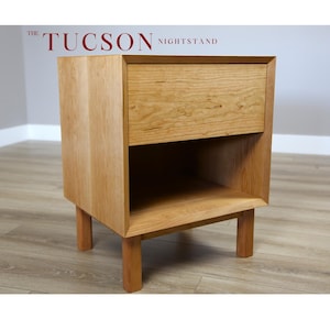 May include: A wooden nightstand with a single drawer and a lower shelf. The nightstand is made of light wood with a natural finish. The text "THE TUCSON NIGHTSTAND" is printed on the top of the nightstand.