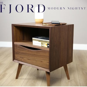 May include: A dark brown wooden nightstand with a drawer and a shelf. The nightstand has a modern design with tapered legs. The nightstand has a lamp, a phone, and a stack of books on top.