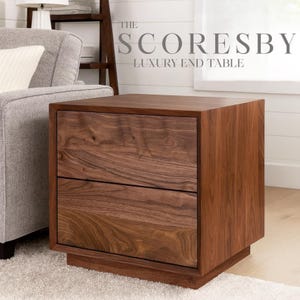 May include: A dark brown wooden end table with two drawers. The table has a modern design with a square shape and a solid base. The text "THE SCORESBY LUXURY END TABLE" is displayed at the top of the image.