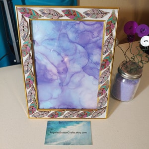 May include: A white picture frame with a gold border and a decorative trim of colorful feathers. The frame is empty and ready to display a photo or artwork.