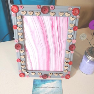 May include: A gray picture frame decorated with pink, red, and white buttons. The buttons have the word "love" written on them. The frame has a pink and white marbled background.