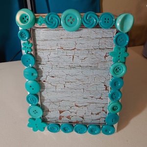 May include: A silver picture frame decorated with turquoise buttons of various shapes and sizes. The frame has a distressed, light blue background.