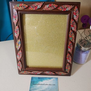 May include: A brown wooden picture frame with a gold glitter background. The frame is decorated with colorful patterned fabric leaves.