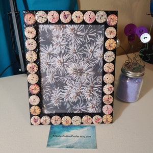 May include: A black picture frame decorated with vintage-style buttons featuring floral and clock designs. The frame is holding a photograph of a snowy evergreen tree.
