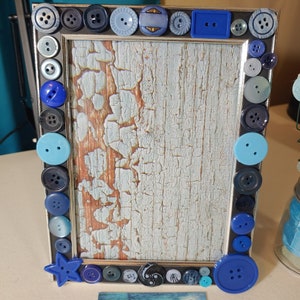 May include: A blue picture frame decorated with blue buttons of various sizes and shapes. The frame has a light blue and white distressed background.
