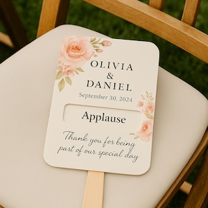 Op de afbeelding: Een bruiloftwaaier met de namen "OLIVIA & DANIEL" en de datum "30 september 2024". De waaier heeft een bloemenontwerp met perzikrozen en de woorden "Applause" en "Thank you for being part of our special day".
