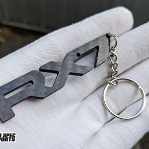 Op de afbeelding: Een zwarte carbon fiber sleutelhanger met het woord "RX7" uitgesneden. De sleutelhanger is bevestigd aan een zilveren sleutelring.