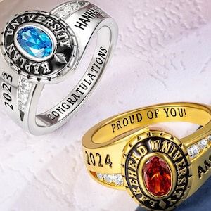 May include: Two graduation rings, one silver and one gold. The silver ring has a blue gemstone and the text "Kaplan University 2023 Congratulations". The gold ring has a red gemstone and the text "Lakehead University 2024 Proud of You!"