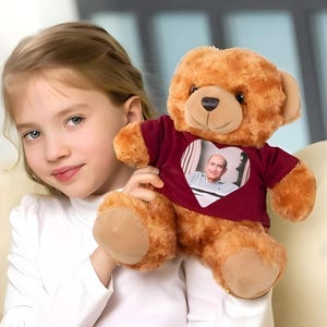 Può includere: Un orsacchiotto di peluche marrone che indossa una maglietta bordeaux con una foto a forma di cuore di un uomo. L'orso è tenuto da una bambina.