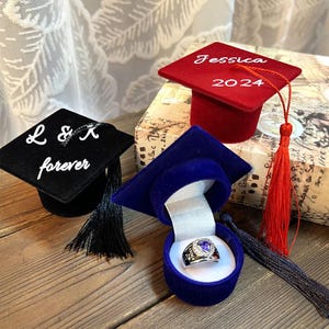 Könnte beinhalten: Drei Ringboxen in Form von Abschlusskappen. Eine ist schwarz mit dem Text "L & K forever", eine ist rot mit dem Text "Jessica 2024" und eine ist blau mit einem Ring darin.