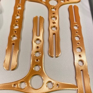 May include: Four gold-coloured metal pieces with holes and a textured surface. The pieces are shaped like a T, a Y, and two straight pieces. The text "Silk Gold" is visible at the top of the image.